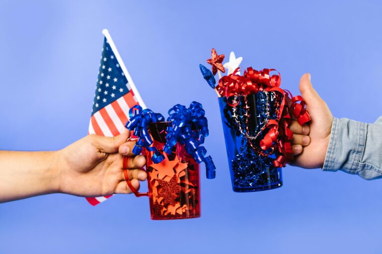 Hands holding decorated cups and American flag on a bright background, symbolizing celebration.