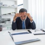 An elderly businessman in a suit appears stressed while sitting at a desk with documents in an office.