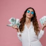 Elegant woman in sunglasses holding euro banknotes against pink background, showcasing confidence and financial empowerment.